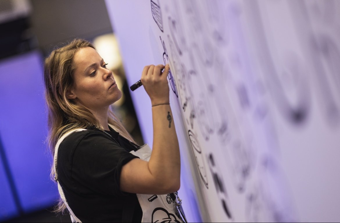 Woman drawing on a large whiteboard with black markers in an indoor setting.