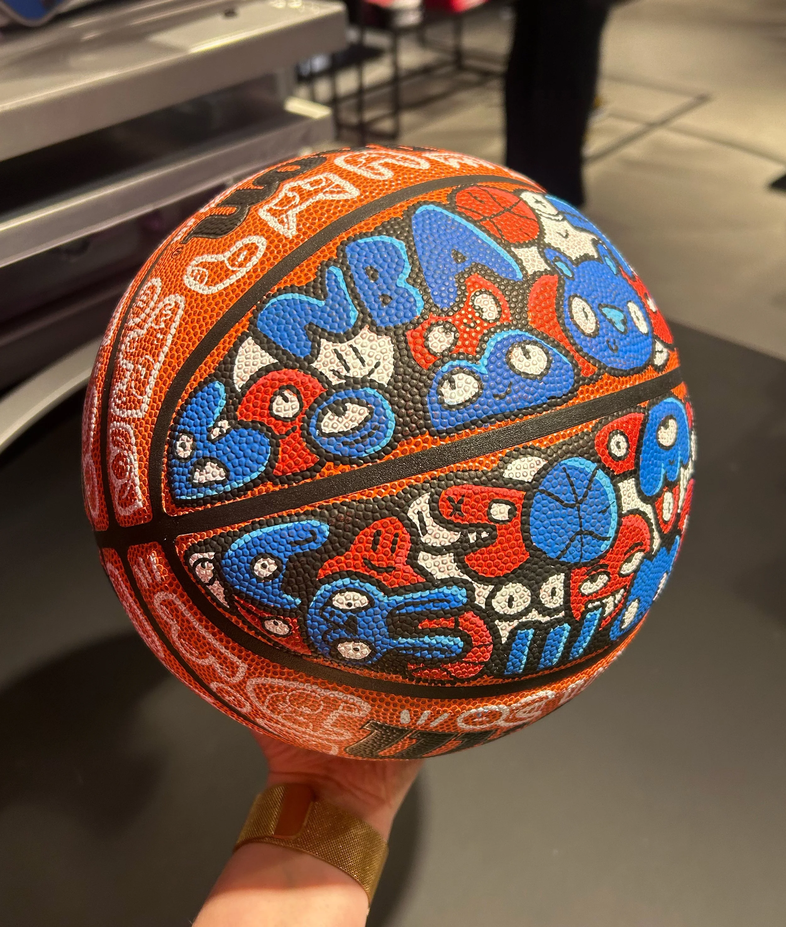 A close-up of a speckled orange and black basketball with colorful graffiti-style text and graphics, being held by a person with a gold wristband in an indoor setting.