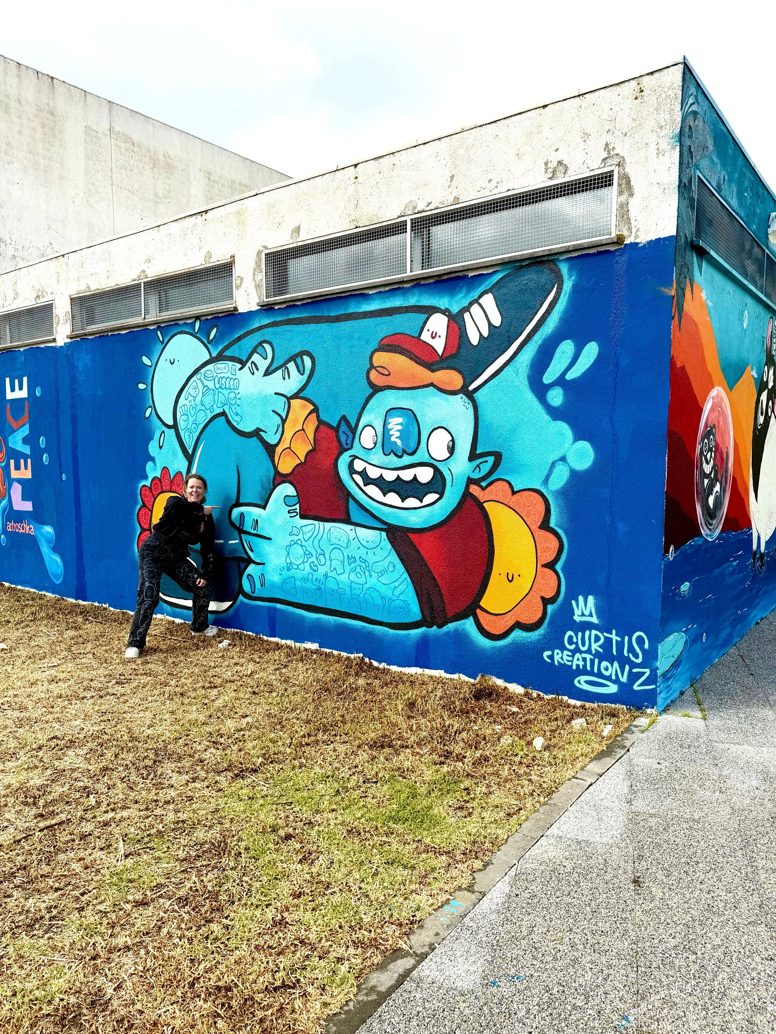 Colorful mural on building wall featuring a blue monster character with a happy expression, holding a watering can, in front of a flower and sun. Person standing in front smiling and pointing at the mural.