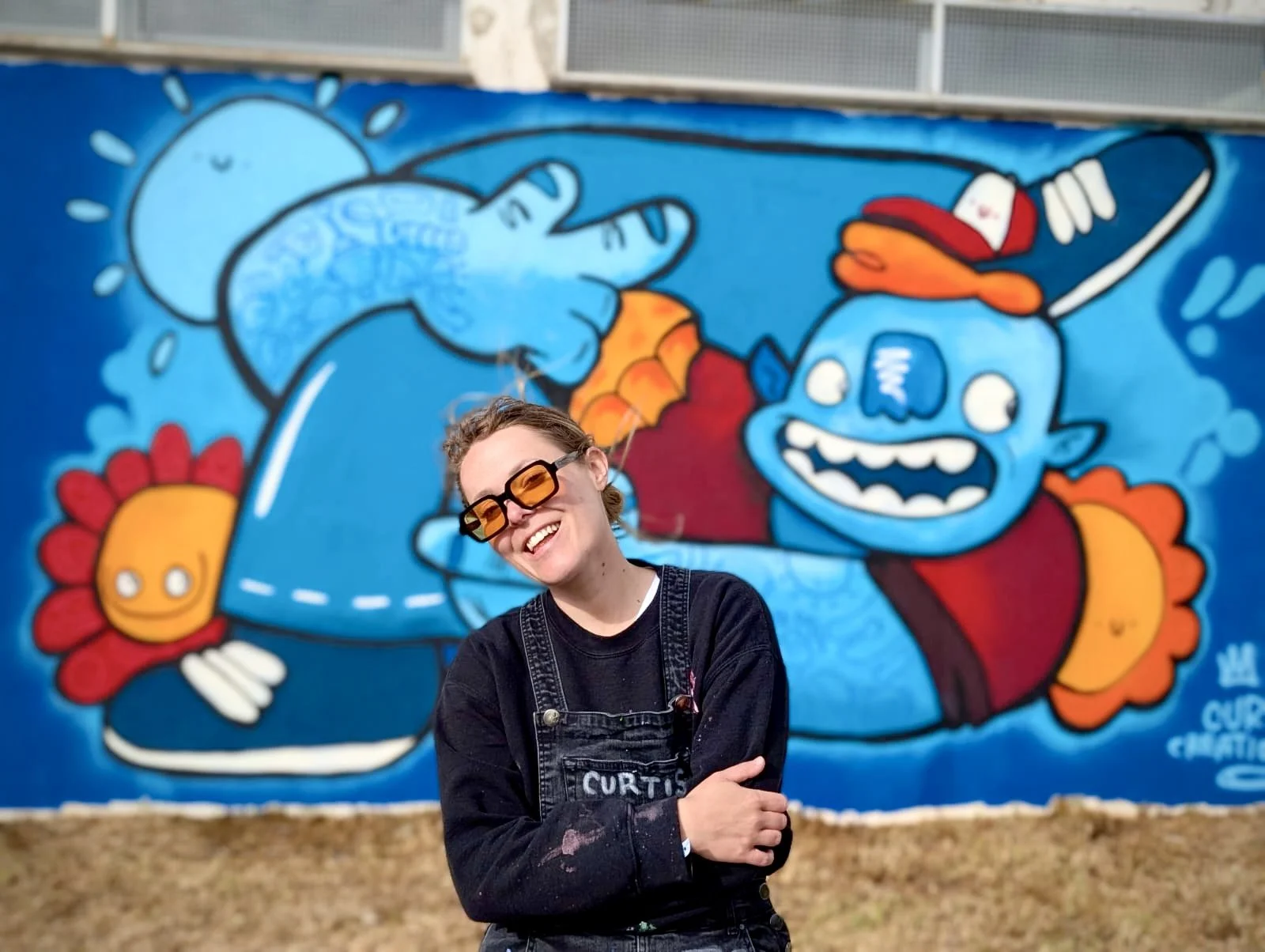 A smiling woman wearing sunglasses and a black shirt with denim overalls stands in front of a colorful mural featuring a cartoonish blue character with a big open mouth, red cap, and flower-like details, smiling and waving.