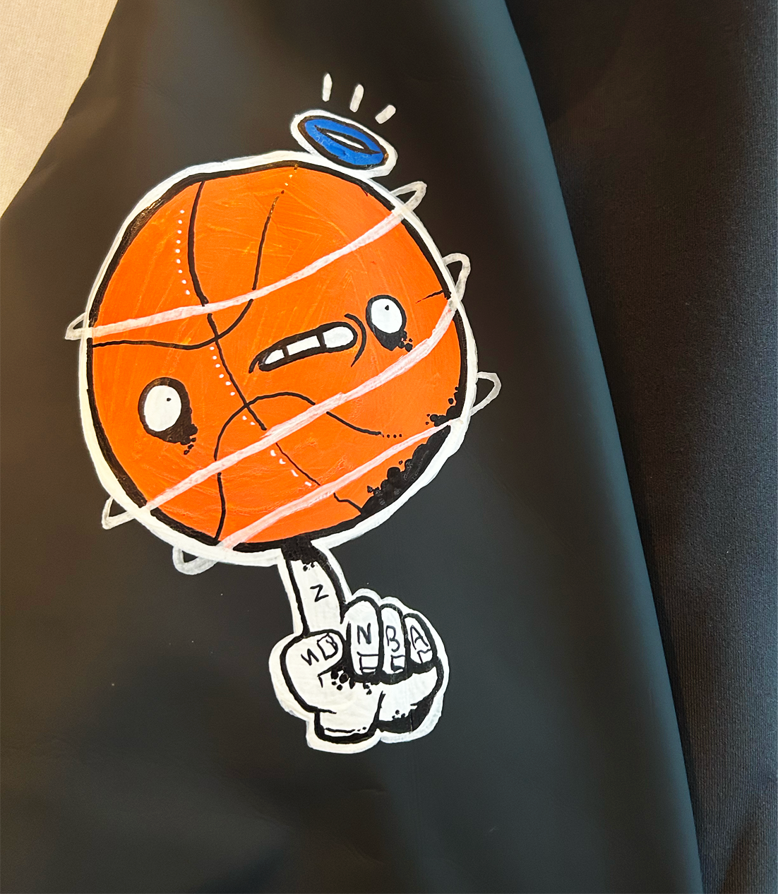 Street art illustration of an orange basketball with an angry face, wearing a blue halo, with white lines and a black outline. The basketball has a hand with a finger pointing up, showing tattooed fingers with phrases.