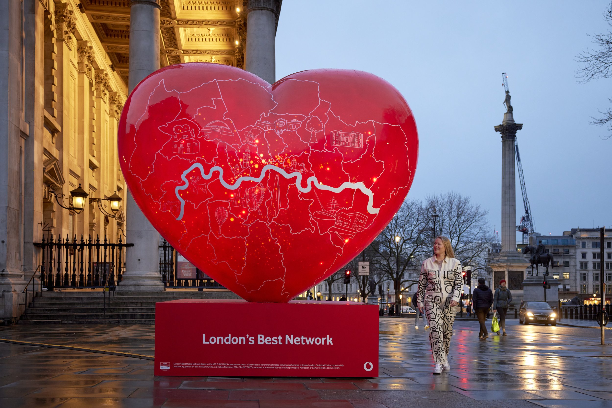 A large red heart-shaped installation with a map of London and icons representing landmarks. The installation has the text 'London's Best Network' at the bottom and is placed outdoors in front of historic buildings at dusk. People are walking nearby.