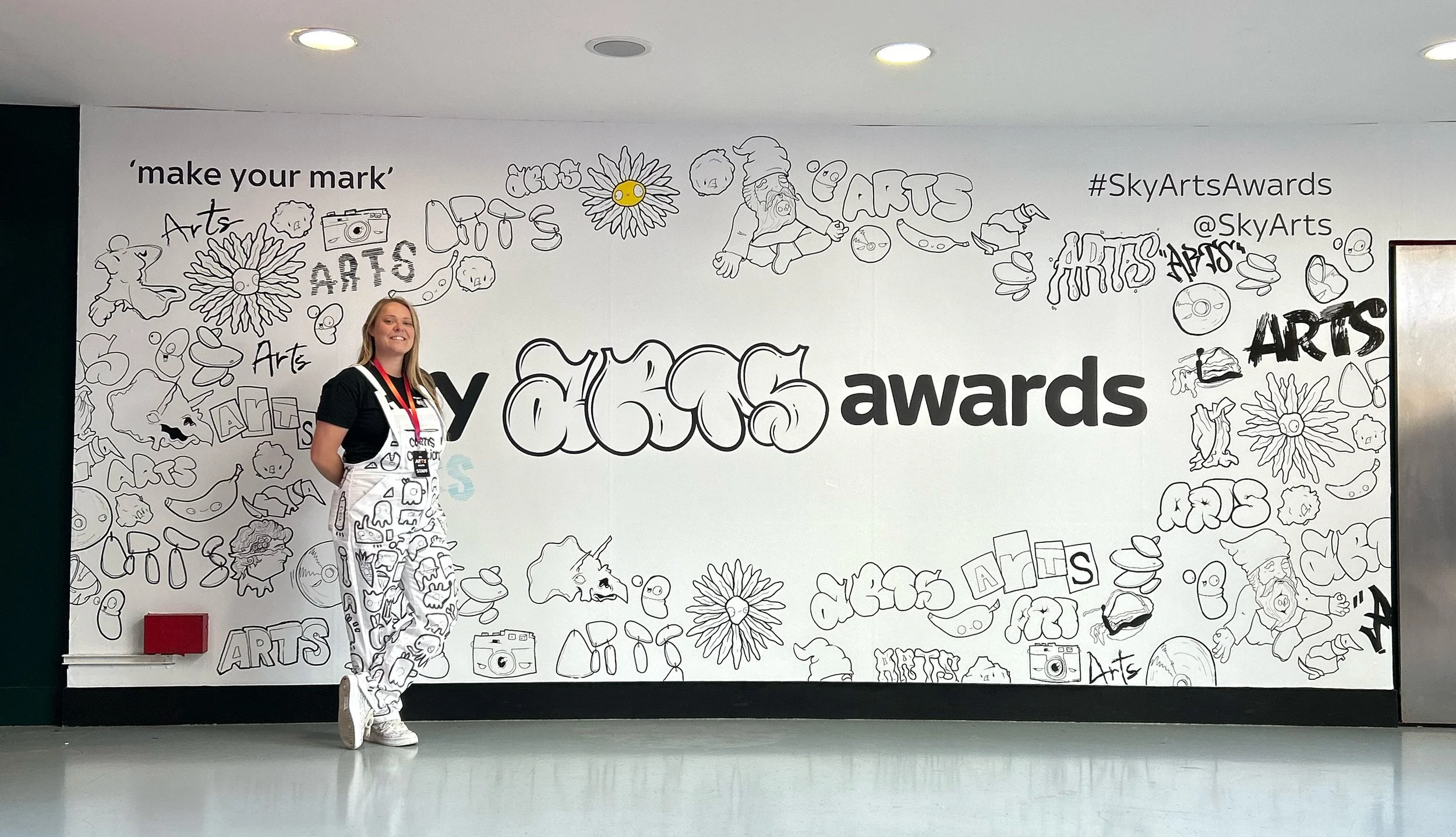 A woman standing in front of a decorative wall at a Sky Arts Awards event. The wall features black and white line drawings of various art-related images and the words 'make your mark' and '#SkyArtsAwards'. The woman is smiling and wearing a black t-s