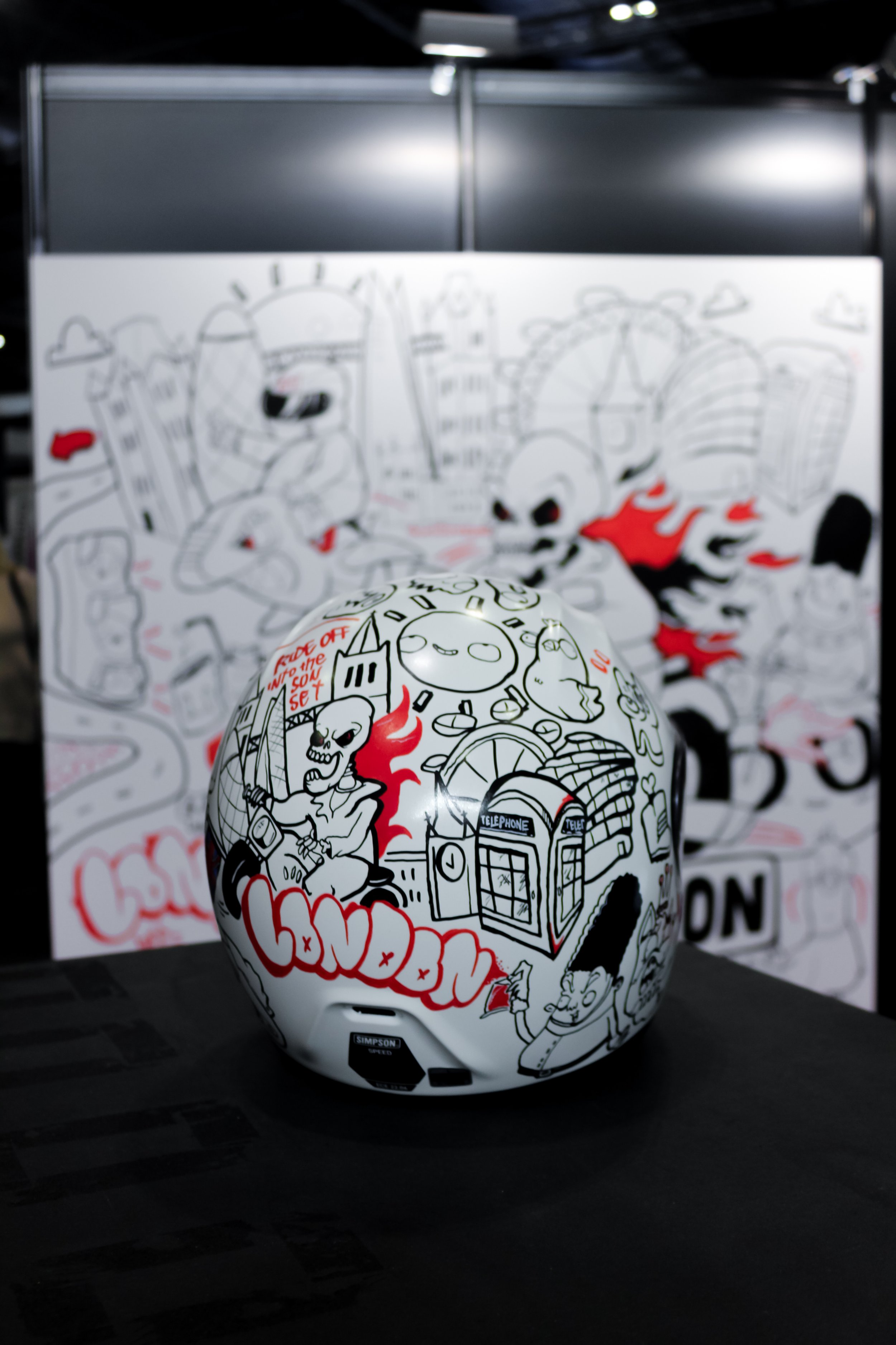 A white helmet featuring black and red street art-style illustrations, including a skeleton, a phone booth, clouds, flames, and various other designs. In the background, there is a white poster with similar black, red, and gray graffiti-style artwork