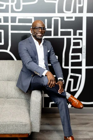 A smiling man in glasses and a blue-grey suit sits on the arm of a sofa in front of a black-and-white street-map mural.