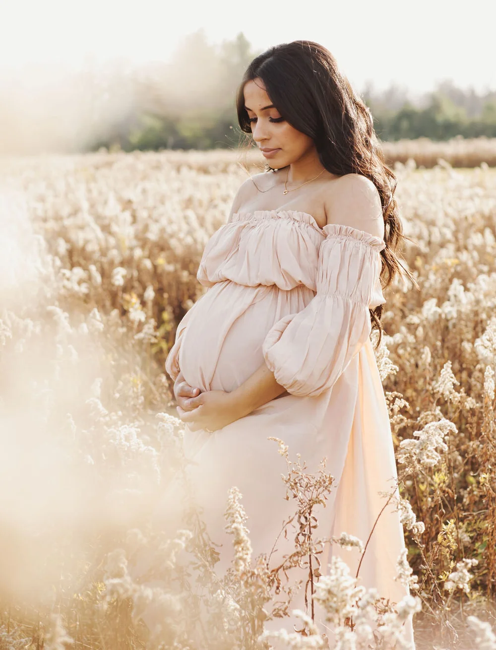 A pregnant woman in a blush off-shoulder dress looks down at her bump, cradling it in both hands, in a hazy field of dried wildflowers.