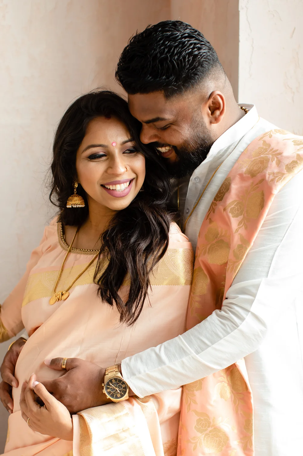 A laughing man hugs his pregnant partner from behind, his hand over hers on her bump, as she smiles with her eyes half closed.