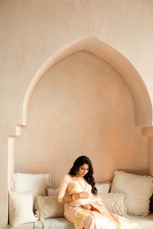 A pregnant woman in a peach silk saree sits on cushions in a tall plaster archway, looking down at her bump with both hands resting on it.