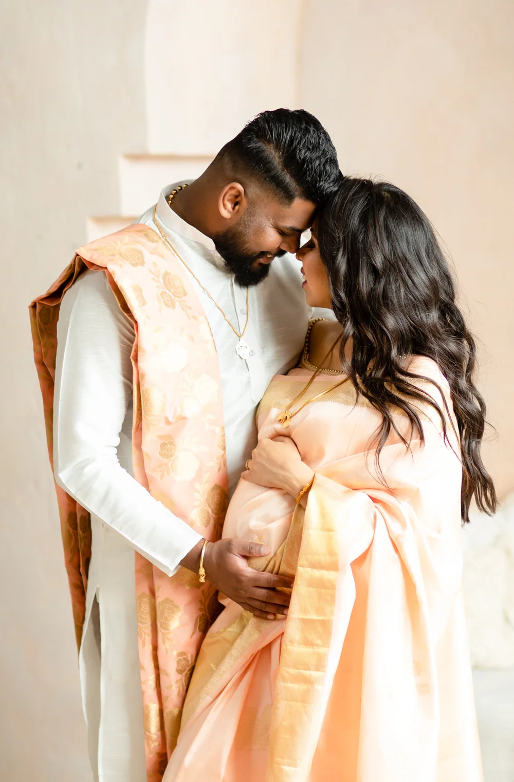 An expectant couple rest their foreheads together, eyes closed, his hand on her bump, in a pale plaster studio.