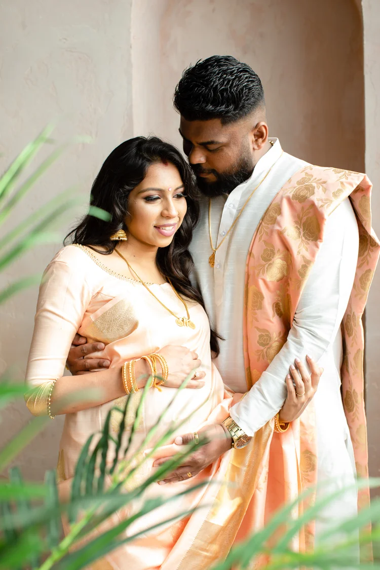 An expectant couple stand in a plaster archway, his arms around her and her hands on her bump, framed by blurred palm leaves.
