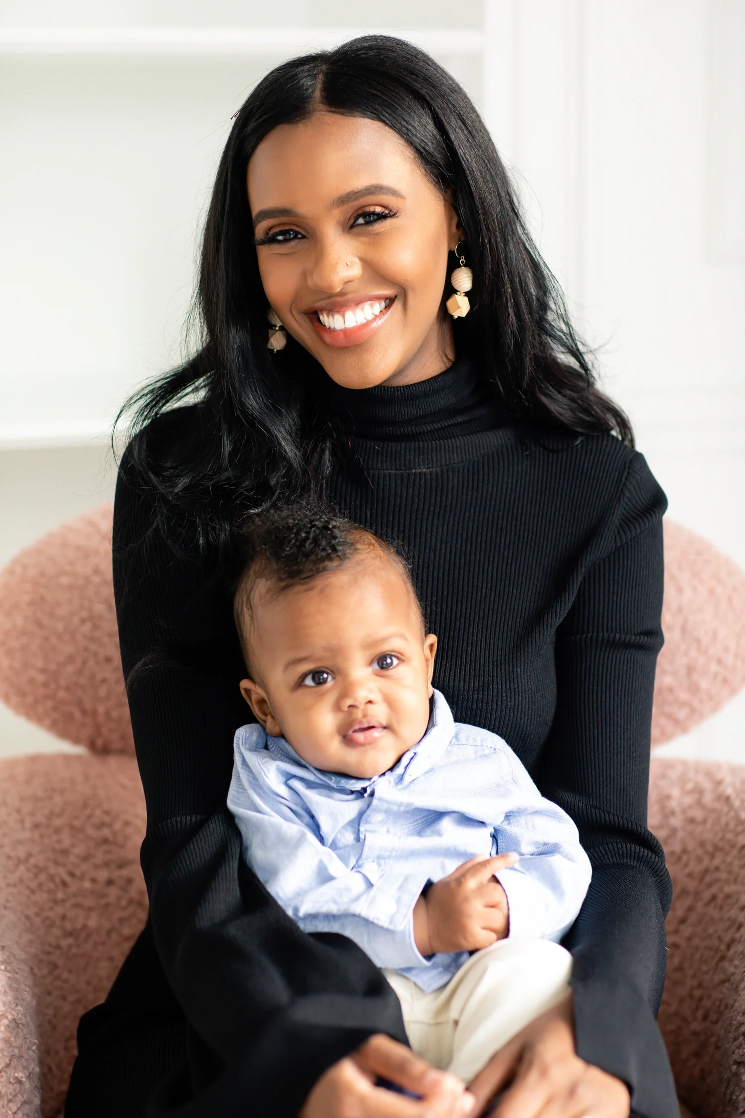 A smiling mother in a black turtleneck holds her baby son on her lap in a blush-pink armchair; both look at the camera.