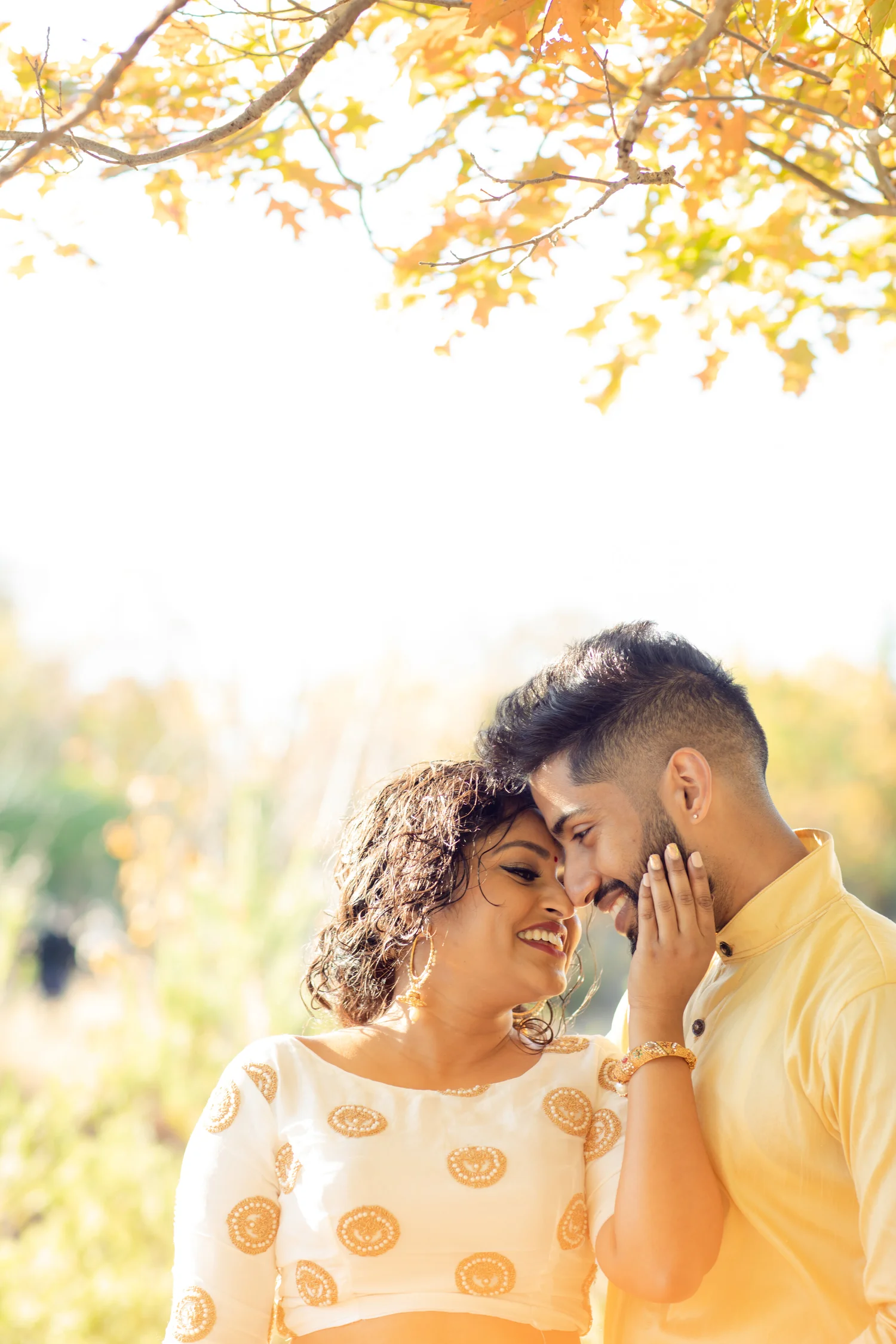 Under autumn leaves, a laughing couple in white-and-gold and pale yellow traditional outfits touch foreheads, her hand on his cheek.