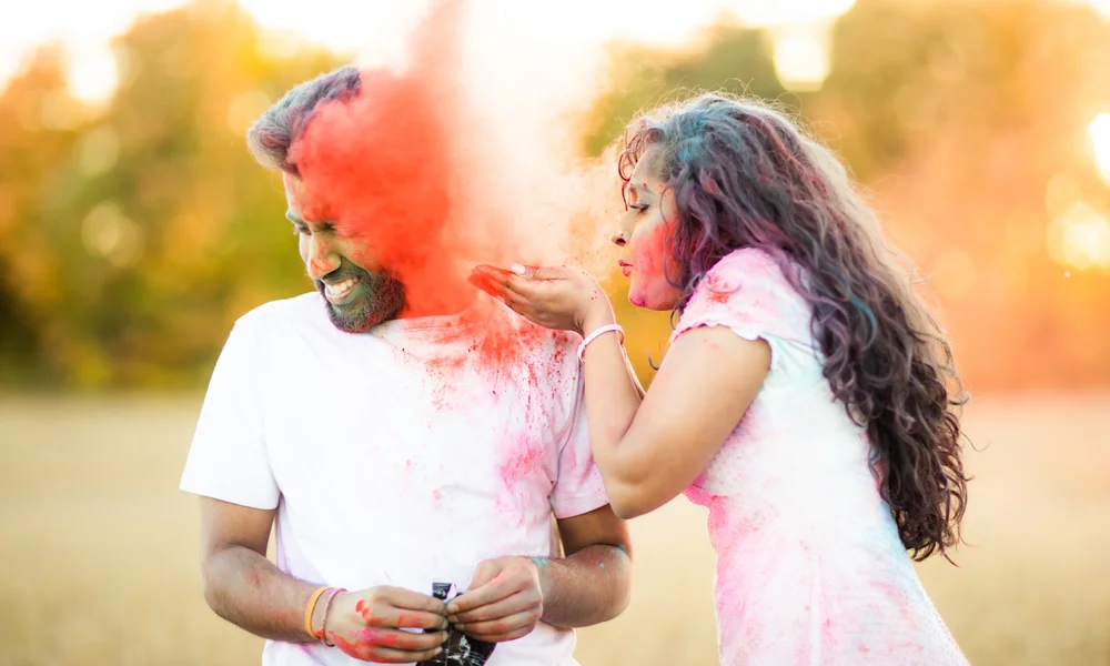 A woman blows a handful of red coloured powder at her laughing partner in a field, both in white already streaked with colour.