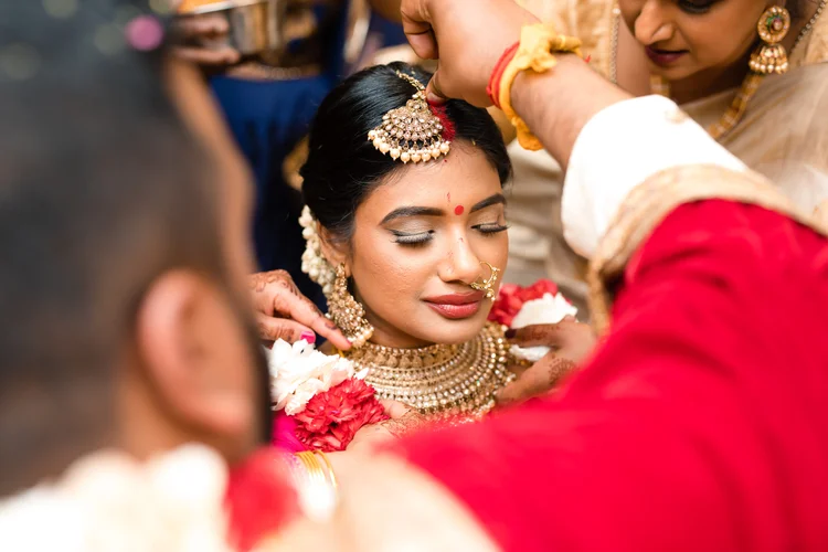 Eyes closed, the bride sits still as the groom's hand marks red kumkum in her hair parting.