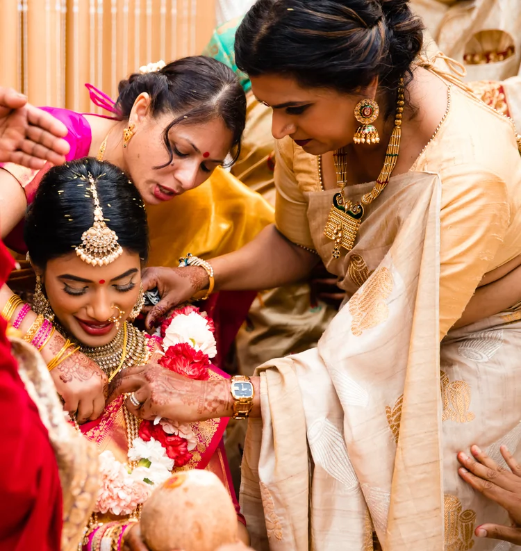 Two women in silk sarees lean over the smiling bride, adjusting her garland as she holds a coconut.