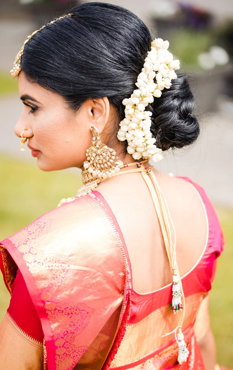 Seen from behind, a bride in a pink and gold silk saree wears jasmine in her low bun and a gold maang tikka.