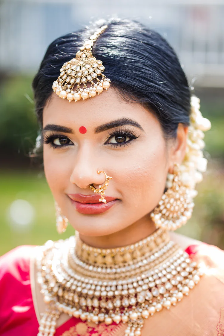 A bride in a pink silk saree and gold bridal jewellery looks straight at the camera with a soft smile.