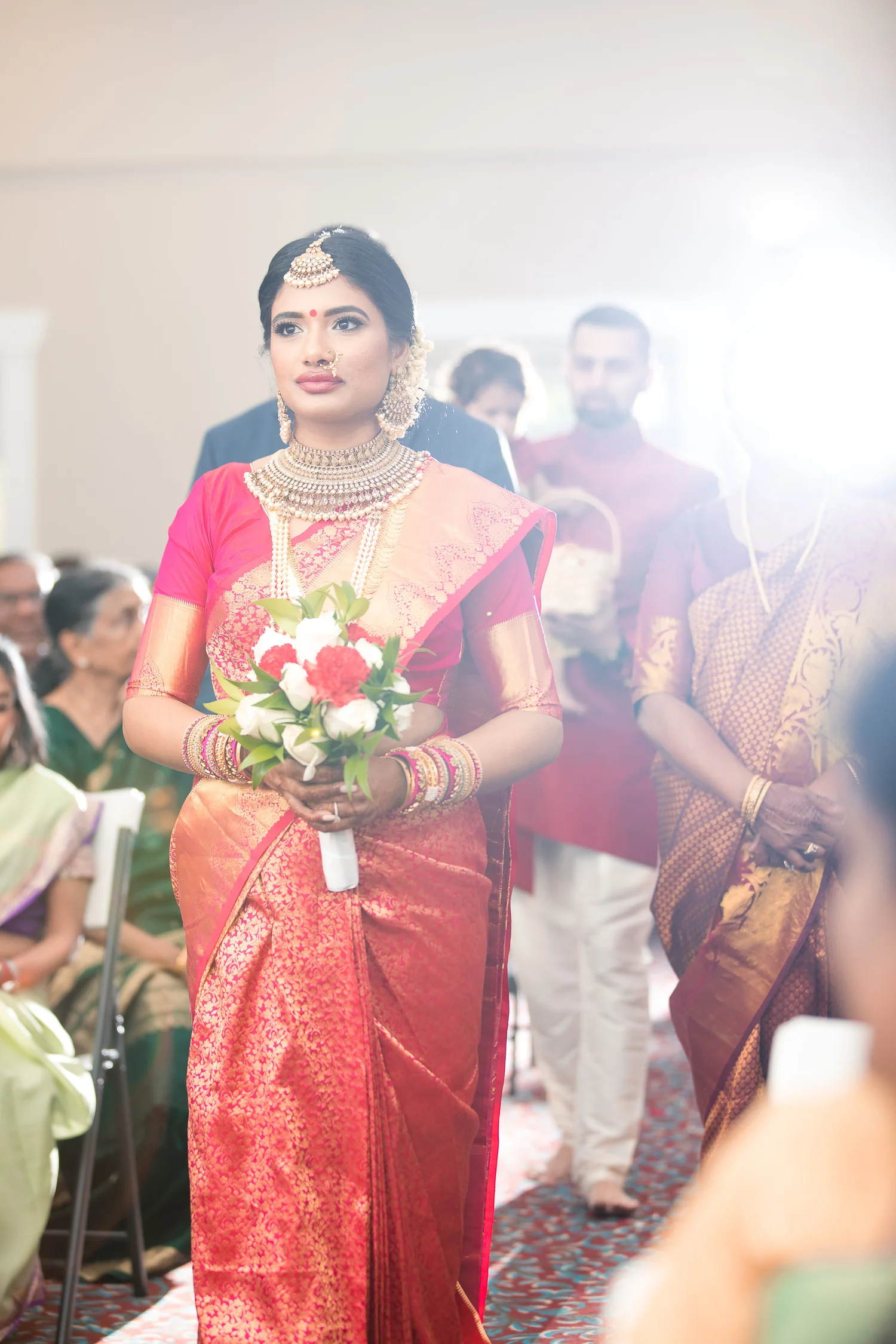 A bride in a red and gold silk saree walks down the aisle holding red and white roses, relatives beside her and guests on both sides.
