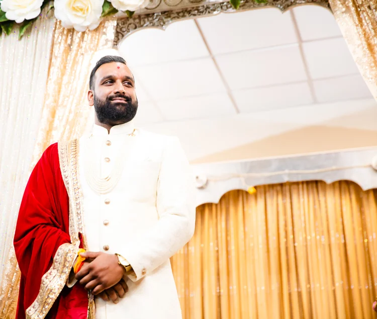 The groom in a cream sherwani and red velvet shawl waits, smiling, under a mandapam hung with gold drapes and white roses.