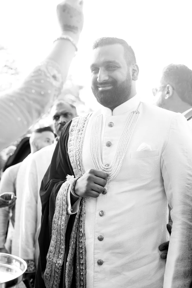 In black and white, the groom smiles in a white sherwani and long pearl necklaces as the men around him cheer.