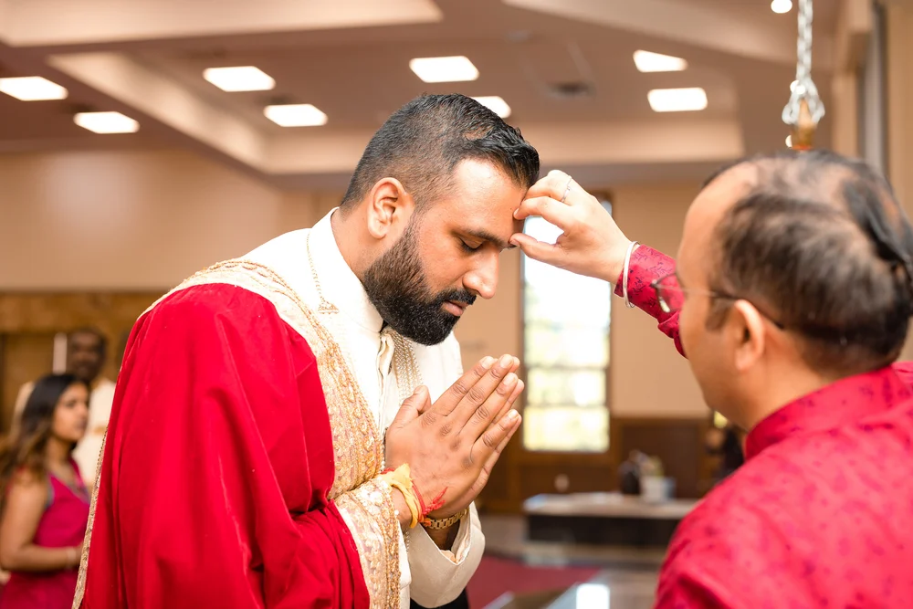 A man in a magenta kurta presses his fingers to the groom's forehead in blessing as the groom bows, eyes closed, palms joined.