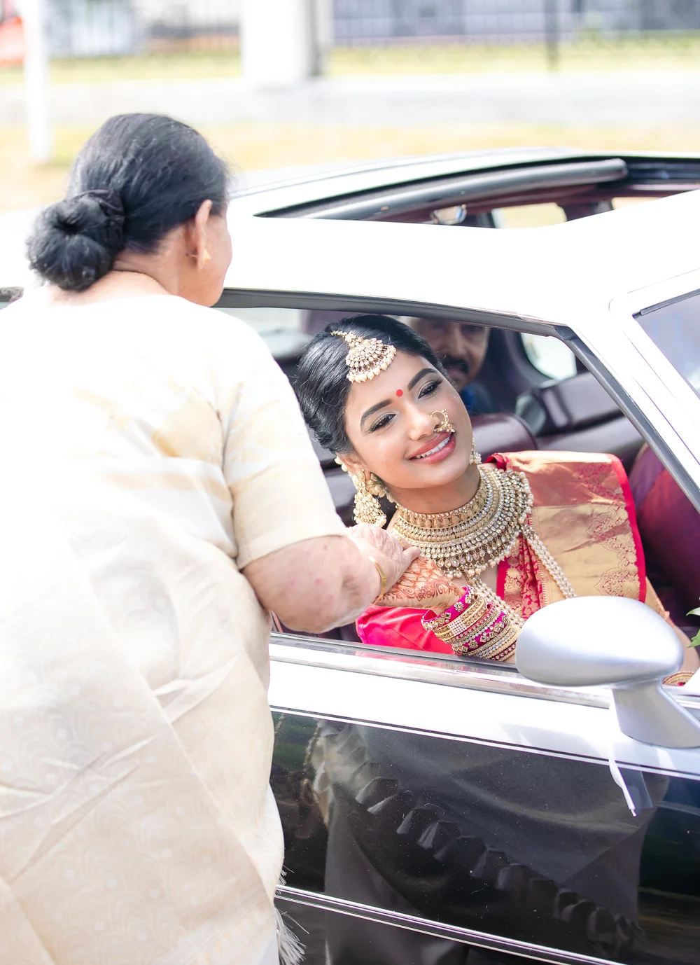 An older woman in a cream saree holds the bride's hennaed hand at the window of a silver car as the bride smiles up at her.