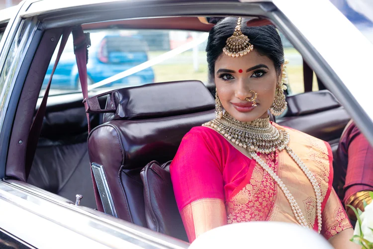 A bride in a pink and gold silk saree and gold bridal jewellery sits in the back seat of a vintage car, smiling at the camera.