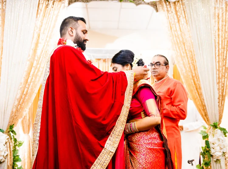 Under a gold-draped mandapam, the groom in a red velvet shawl touches the bride's hairline as a priest in orange looks on.
