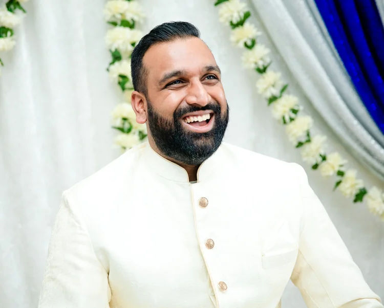 A bearded groom in a cream sherwani laughs beside strands of white jasmine and silver and royal-blue drapes.