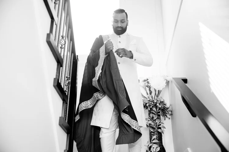 In black and white, a groom in a white sherwani comes down the stairs settling a dark embroidered shawl over his arm.