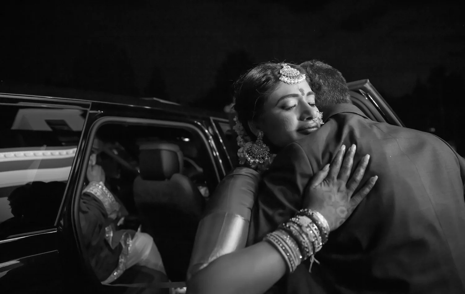 In black and white at night, the bride closes her eyes as she hugs a man in a dark suit beside an open car door.