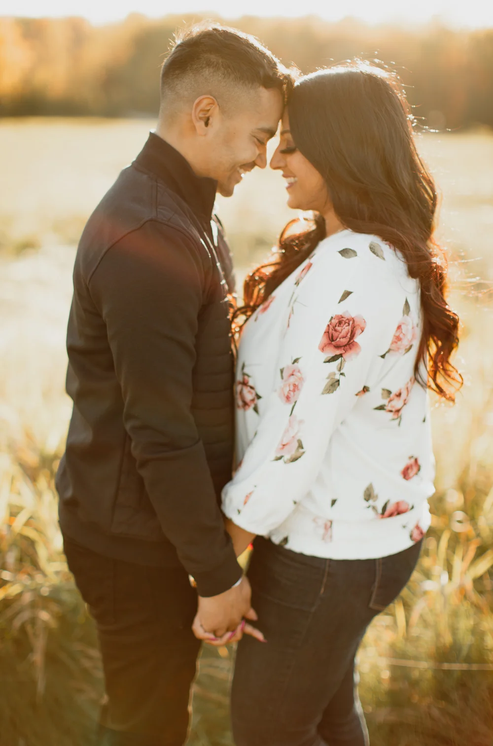 Backlit in a golden field, a couple stand forehead to forehead, holding hands and smiling.