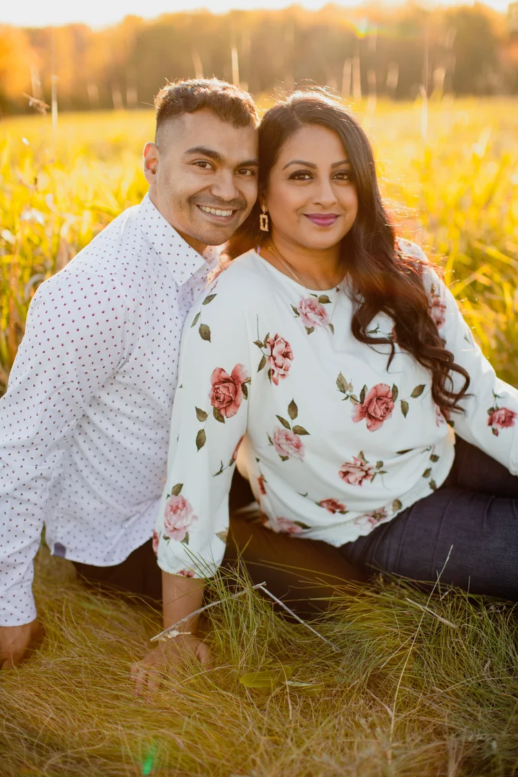 A smiling couple sit close together in a golden grass field, he in a white dotted shirt and she in a rose-print blouse.