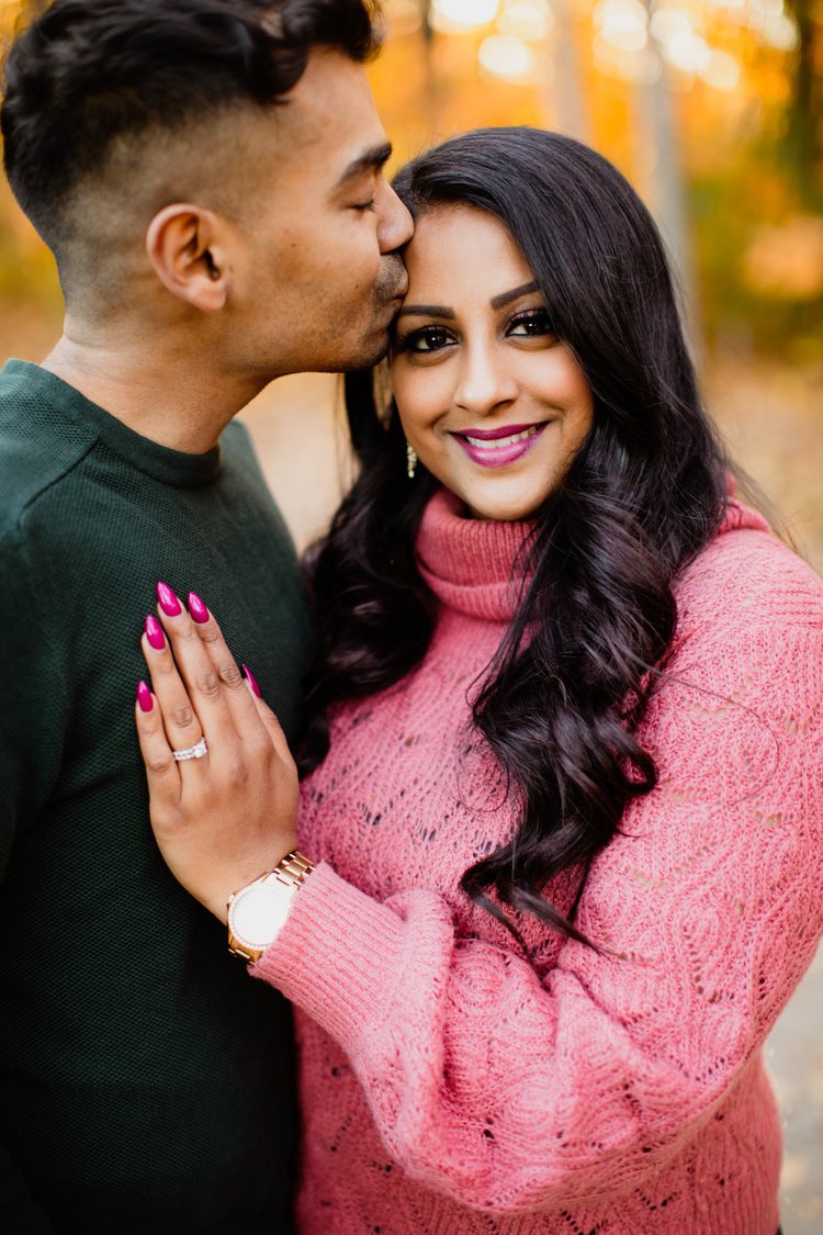 A man in a green sweater kisses the temple of a smiling woman in a pink turtleneck, her ringed hand on his chest, autumn trees behind.