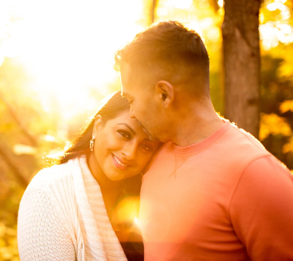 In bright low sun among autumn trees, a smiling woman leans her head on her partner's chest as he kisses her hair.
