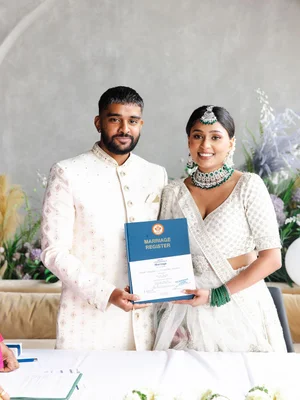 A smiling couple in ivory, the bride in emerald jewellery, hold up a Marriage Register at a table decorated with white flowers.