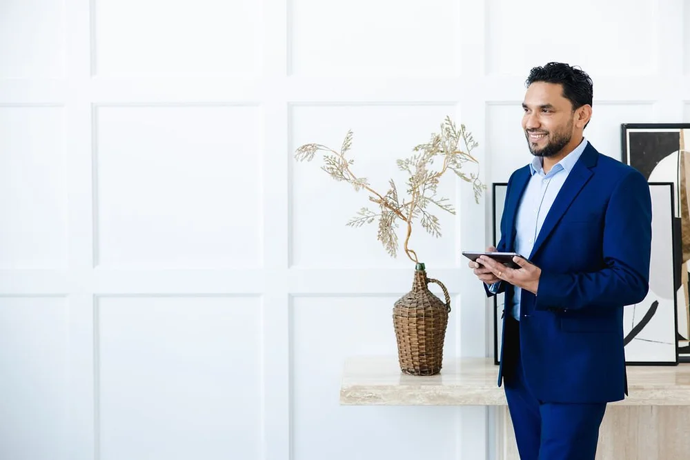 A man in a royal-blue suit smiles down at his phone beside a stone console with a vase of dried branches, in a white panelled room.