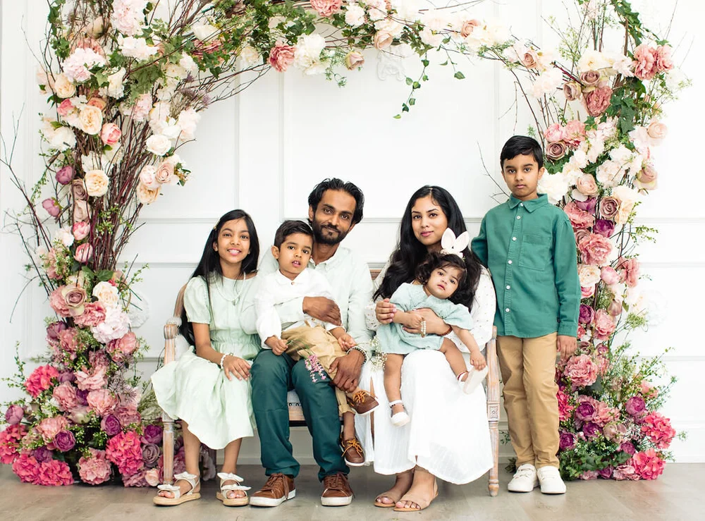 A mother and father with their four children, from a baby in a bunny-ears headband to a boy of about nine, smile together under an arch of pink and cream roses.