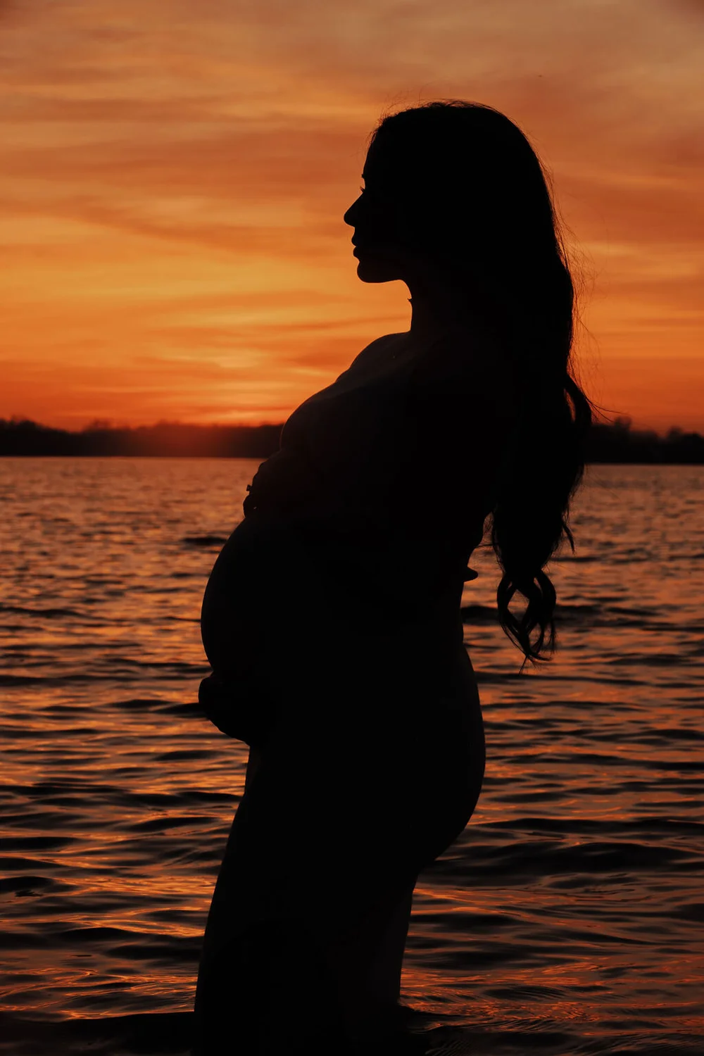 Silhouette of a pregnant woman in profile, standing in a lake against an orange sunset sky.