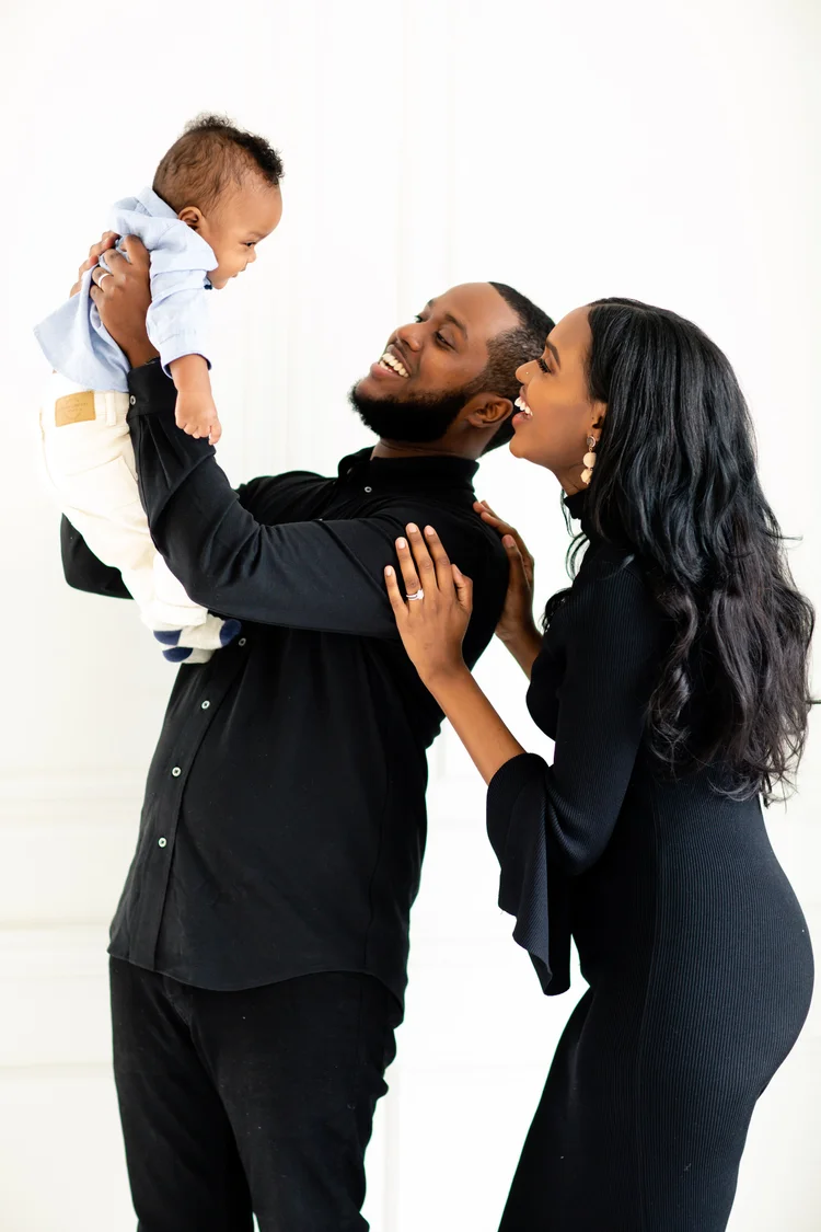 A father lifts his laughing baby son into the air while the mother leans on his shoulder, laughing, in a bright white studio.