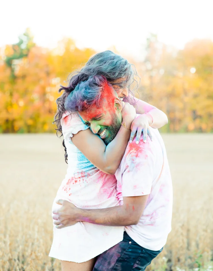 A laughing couple covered in bright coloured powder embrace in a field in front of autumn trees.