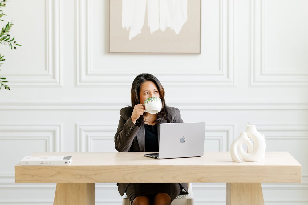 A businesswoman in a charcoal blazer sips from a mug at a pale oak desk with her laptop open, in a bright white panelled room.
