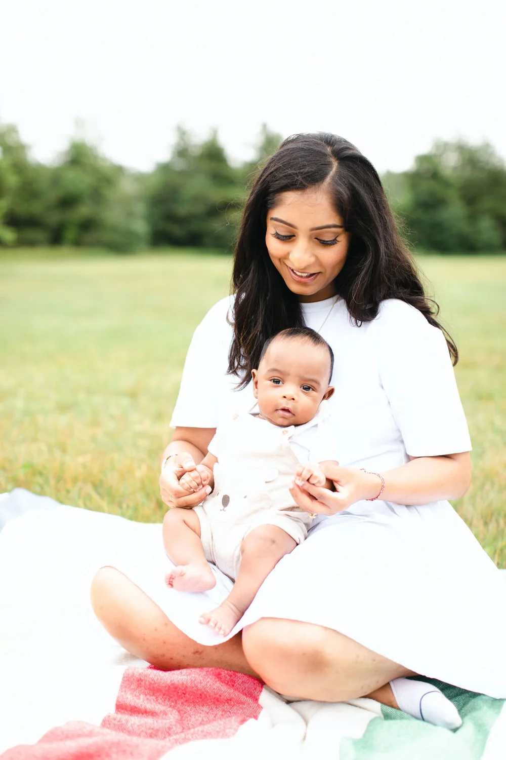 A mother sits cross-legged on a striped blanket in a meadow, smiling down at her baby son on her lap.