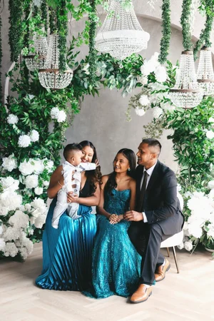 A family of four in formal teal and black sit beneath an arch of white hydrangeas and crystal chandeliers, the mother holding her baby son.