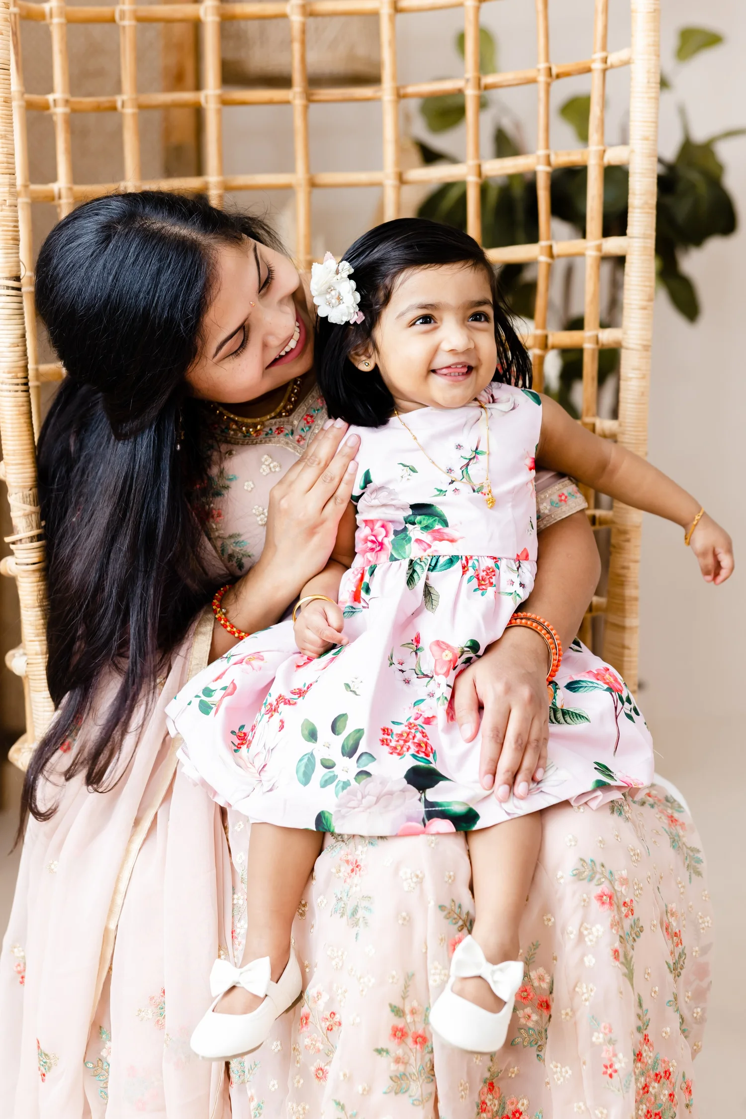A smiling mother holds her toddler daughter on her lap in a rattan chair, the two in matching pink floral outfits.