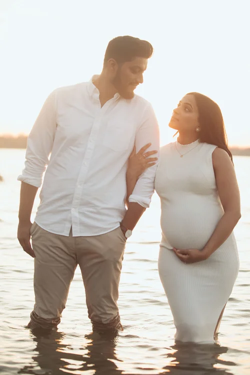 An expectant couple stand in a lake and gaze at each other, her hand on his arm and the other under her bump.