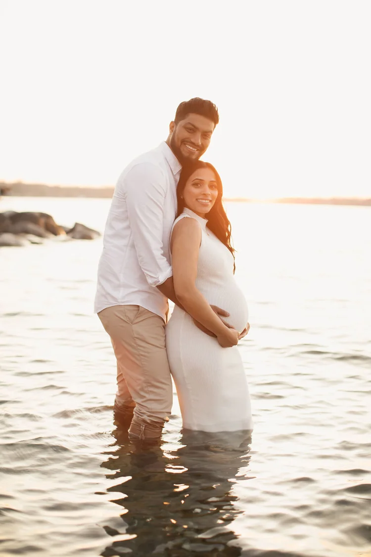 An expectant couple stand knee-deep in a lake, he hugs her from behind with his hands on her bump, both smiling at the camera.