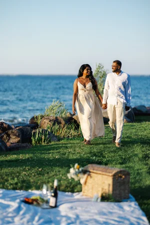 A couple in cream and white walk hand in hand across a lakeside lawn, a picnic blanket and hamper in the foreground.