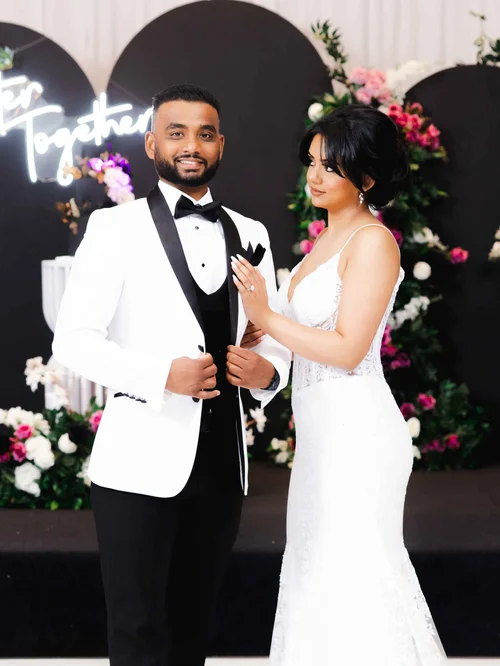 A groom in a white tuxedo jacket and a bride in a white lace gown pose before black arches and pink flowers at their reception.