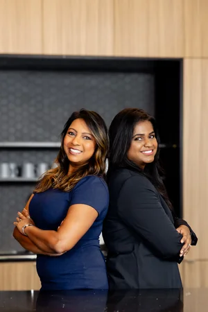 Two businesswomen stand back to back with arms crossed, smiling, in an office kitchen.